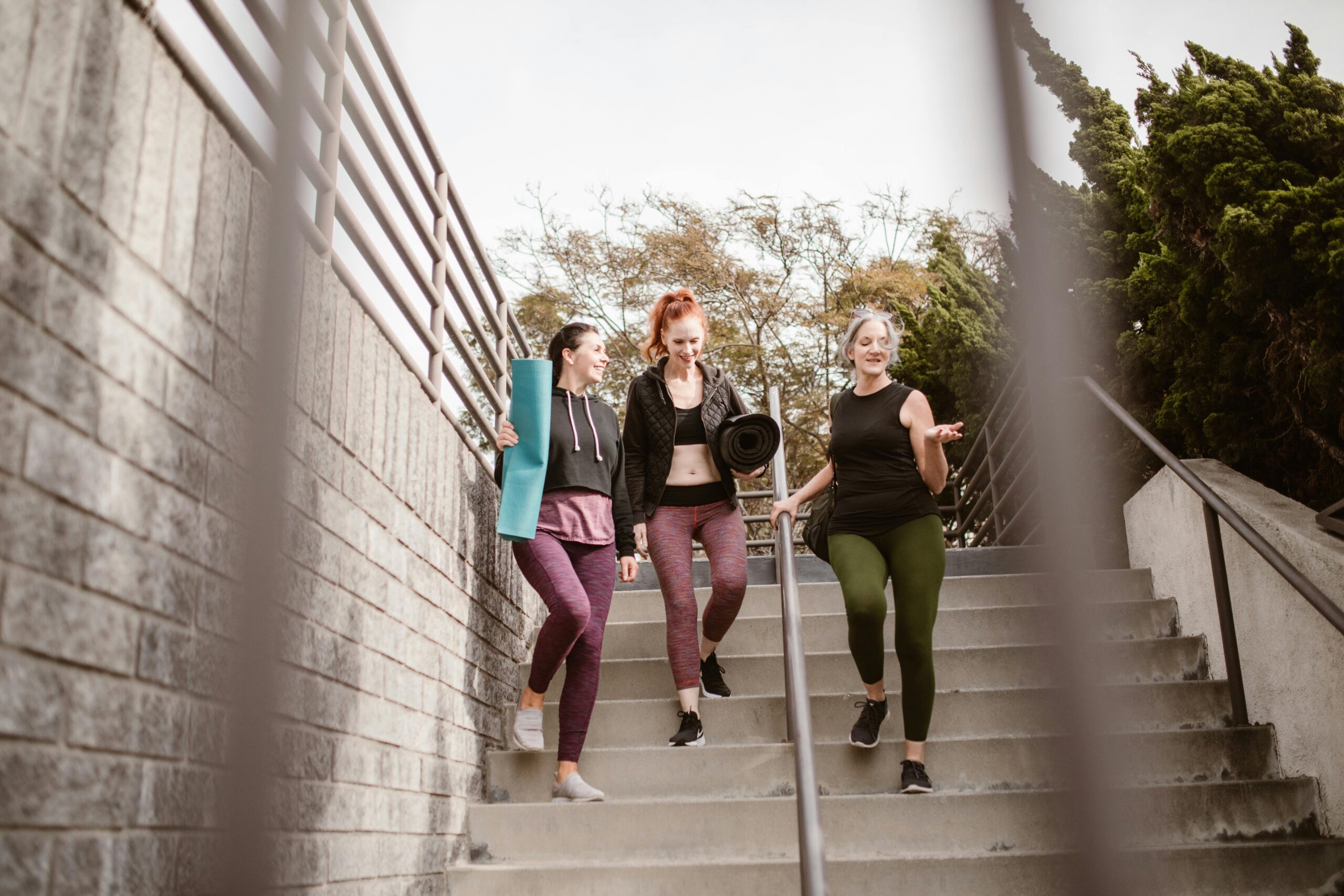 Three women walking down steps outdoors, carrying yoga mats, enjoying a fitness conversation.